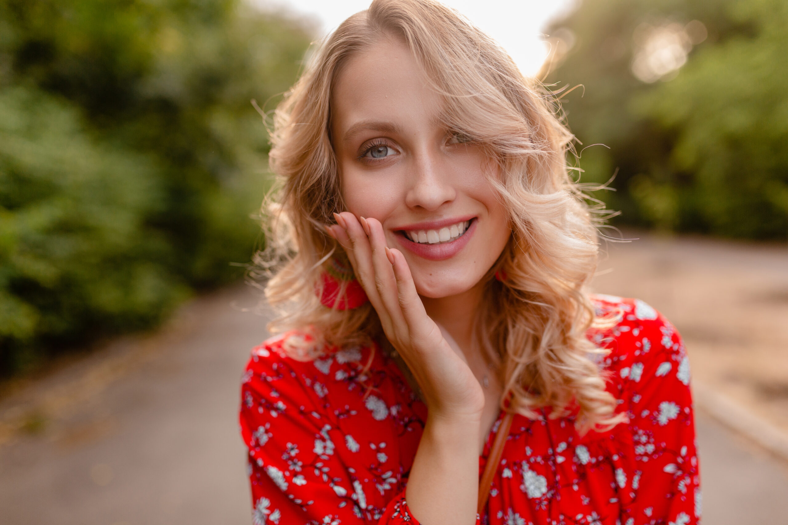 portrait of attractive stylish blond smiling woman in red blouse summer fashion outfit in park boho style wearing earrings smiling white teeth